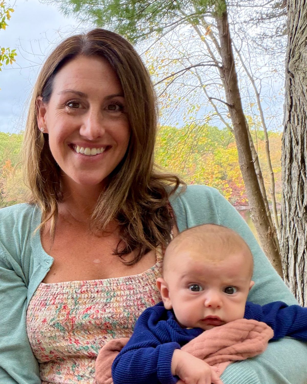 Ashley smiling outdoors among autumn trees, holding her baby